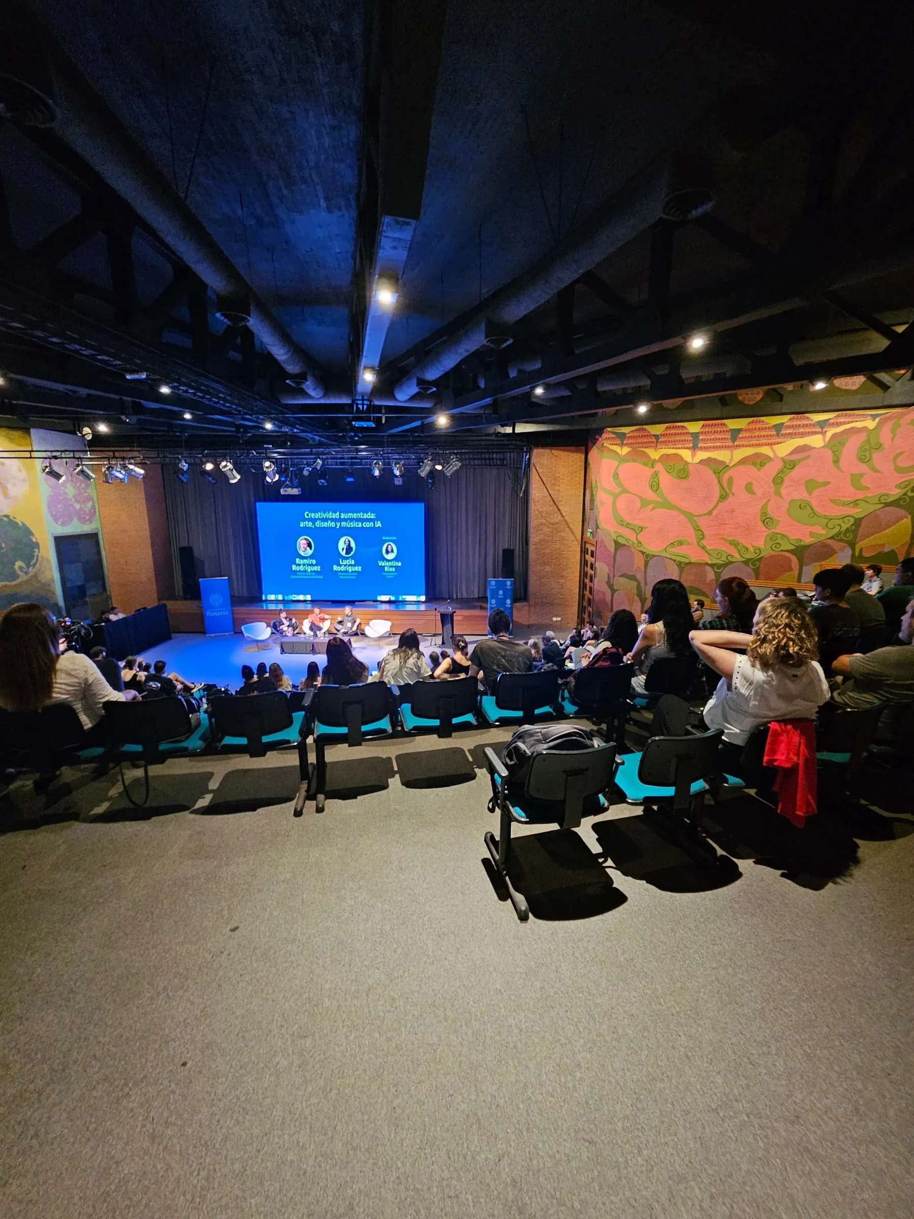 Auditorio con butacas turquesa y público sentado frente a un escenario con un panel de tres personas y una pantalla azul.