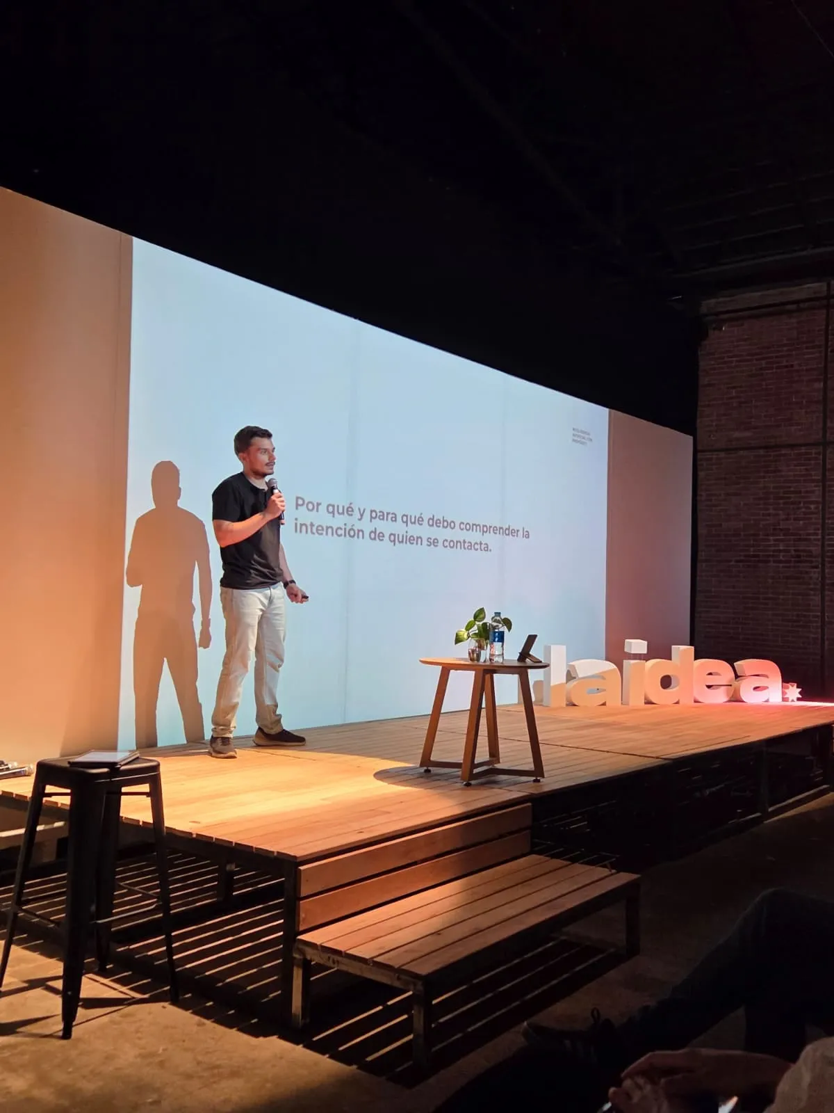 Fran Blasich con micrófono en un escenario de madera, junto a una pantalla con una diapositiva sobre la intención de quien se contacta.